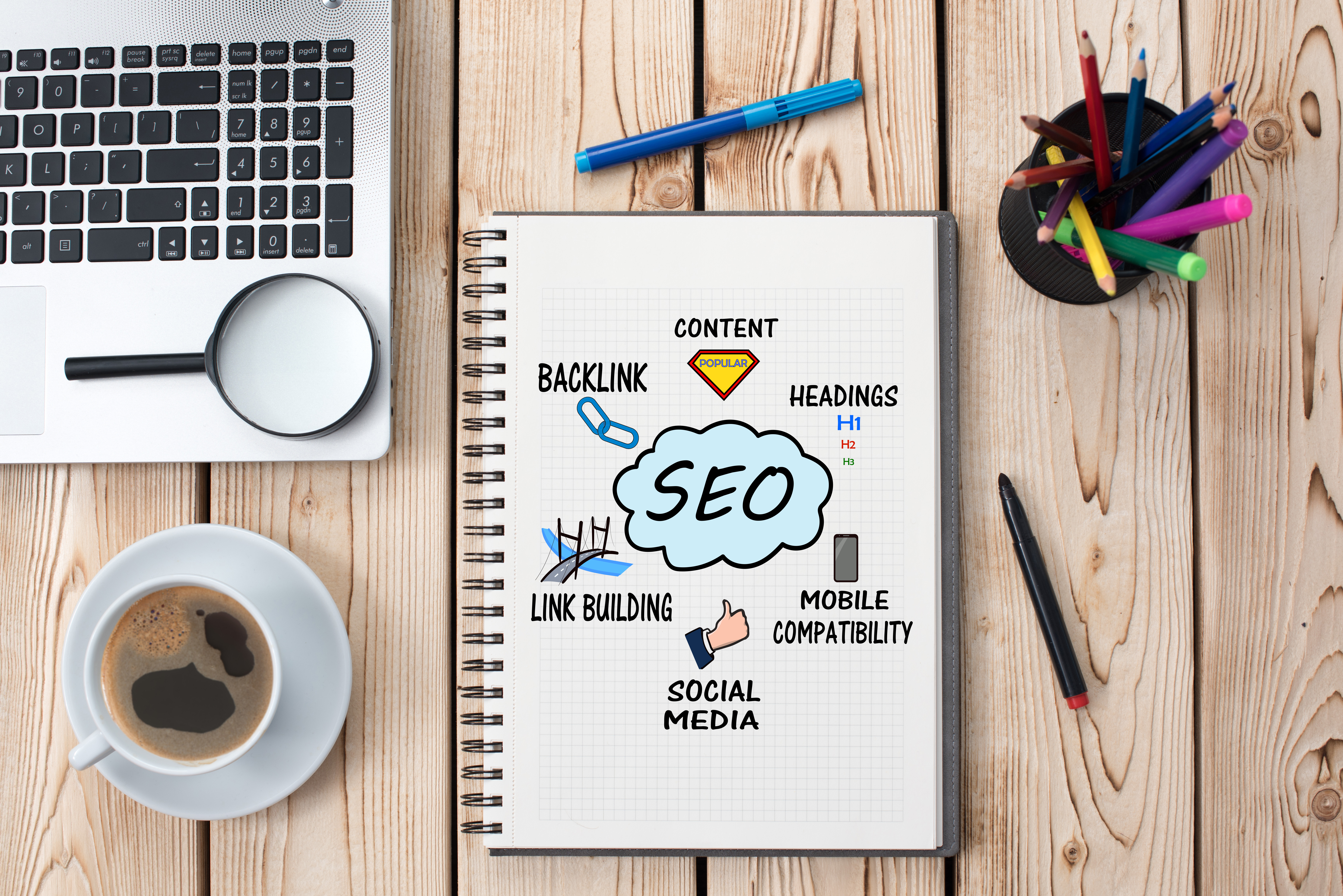 This is a picture of a wooden desk that has items on it. Part of a laptop's keyboard is in view with a magnifying glass situated on top of the laptop right hand lower corner. 
                        There is a saucer and a coffee mug with black coffee in it. 
                        A white note pad with the a world cloud, centered in the middle is the word SEO and around it, content, headings, backlink, link building, mobile compatibility and social media. 
                        Additionally there is a black container with color pencils and markers on the wooden desk. A few pencils and markers are on the wooden desk as well. 
                        